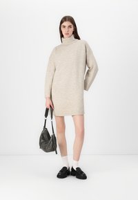 Beige strikket turtleneckkjole med lange ermer, rett snitt, parret med svarte loafers og en grå crossover-veske. Glatt tekstur.