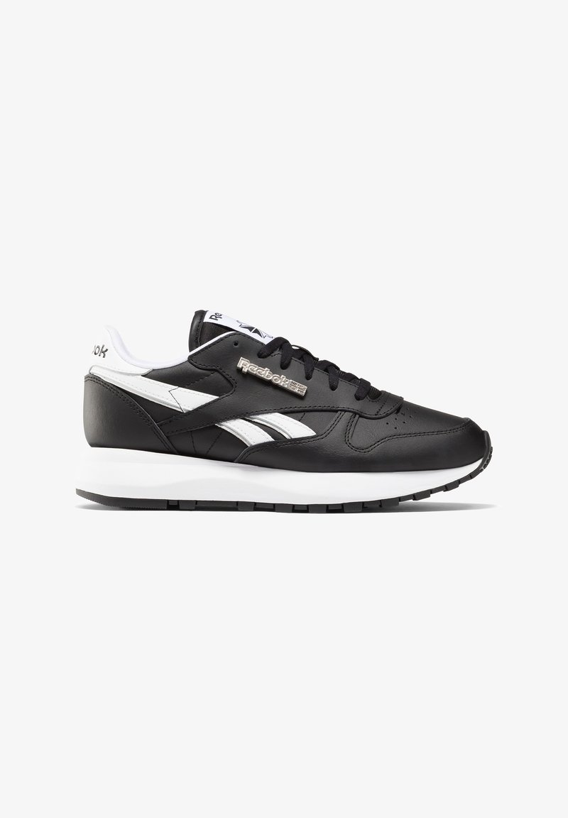 Black leather sneaker with white accents, featuring a padded collar, perforated toe box, and textured rubber sole. Reebok logo is metallic.