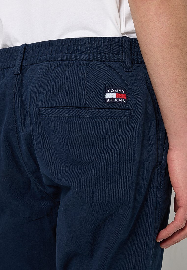 Granatowe bawełniane spodnie z elastycznym pasem, z tylną kieszenią i małym haftowanym logo "TOMMY JEANS" w kolorze czerwonym i białym.