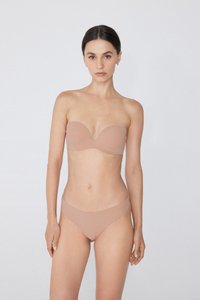 Kobieta z ciemnymi włosami w cielistym biustonoszu bez ramiączek i pasującym gładkim komplecie bielizny, stojąca na tle jasnym.