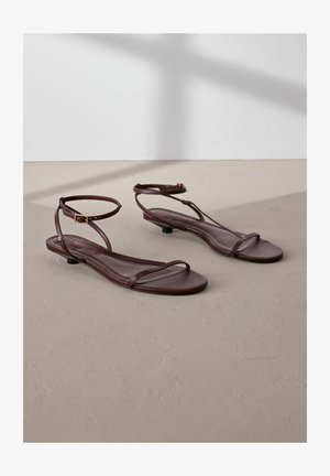 Brązowe skórzane sandały z cienkimi paskami, zapięciem na kostce, płaską podeszwą i minimalistycznym designem. Gładka faktura bez widocznych wzorów.