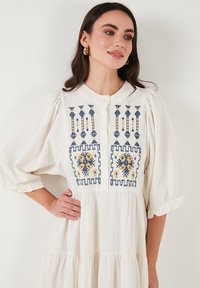 White dress with embroidered navy and yellow floral patterns on the bodice, featuring puff sleeves and a button placket. Soft fabric texture.