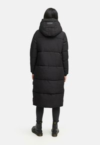 Sort puffersjakke med hætte, prydet med et kviltet design, knælangt snit og lynlås lukning. Bæres over mørke stramme jeans og støvler.