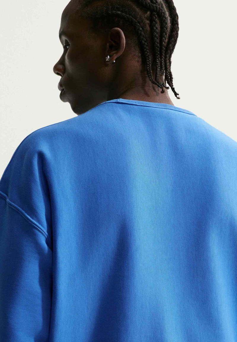 Seitenprofil einer Person mit Cornrows, die einen blauen Pullover trägt und nach links vor einem einfachen weißen Hintergrund schaut.