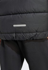 Doudoune noire gonflante avec une texture lisse, présentant une couture visible et une bande réfléchissante horizontale près de l'ourlet.