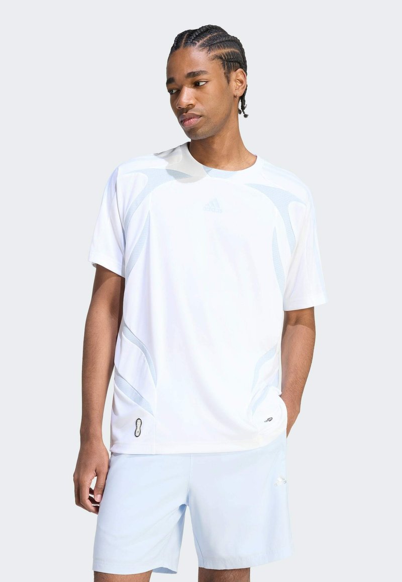 Jeune homme aux cheveux tressés portant un maillot de sport blanc Adidas et un short bleu clair, debout avec une main dans la poche.