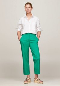 Γυναίκα όρθια με τα χέρια στις τσέπες, φορώντας λευκό πουκάμισο με κουμπιά, πράσινο cropped παντελόνι και μπεζ σανδάλια σε ουδέτερο φόντο.