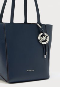 Modra usnjena torba tote z oblikovano zasnovo, ki vključuje srebrno okovje in obesek z logotipom. Gladek tekstura in kontrastni šivi.
