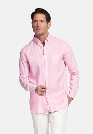 Man met kort bruin haar, gekleed in een lichtroze overhemd met lange mouwen en witte broek, staande tegen een effen achtergrond.