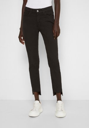 Modelo vistiendo jeans negros ajustados hasta el tobillo con dobladillos sin rematar, combinados con zapatillas blancas y una camiseta blanca sobre un fondo liso.