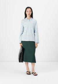 Lyseblå og hvit stripete langermet polo-skjorte med krage, kombinert med et grønt skjørt, bærende en svart tote-bag, iført flip-flops.