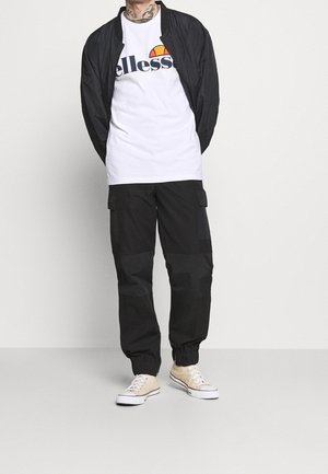 Sort bomberjakke over en hvid t-shirt med en markant logo, kombineret med afslappede sorte cargobukser. Lyse sneakers fuldender looket.