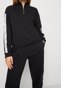 Svart zip-up sweatshirt med hög krage, tillverkad av mjukt tyg, med vit logotape längs ärmarna och en avslappnad passform.