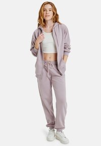 Lavendel zip-up hoodie met zijzakken, gecombineerd met bijpassende joggers. Witte cropped top eronder en chunky witte sneakers. Zachte stoffen textuur.