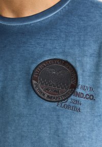 Camiseta de algodón con un degradado azul y un parche negro bordado en forma de círculo que lleva el texto "International Speedway" y "Daytona". Mangas cortas.
