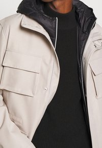 Beige ytterjacka med svart huva, med två stora framfickor och dragkedjestängning. Bärs över en strukturerad svart tröja.