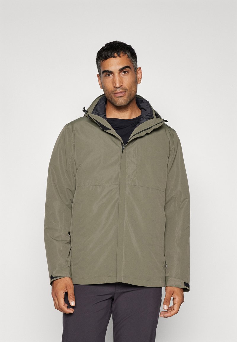 Olijfgroene waterdichte jas met capuchon; voorzien van een ritssluiting, textuurstof en elastische manchetten. Draagt over een zwarte top.