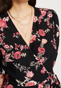 Robe portefeuille noire à fleurs avec des fleurs roses et crème, présentant un décolleté en V profond et de délicates colliers en or superposés. Tissu doux et fluide.