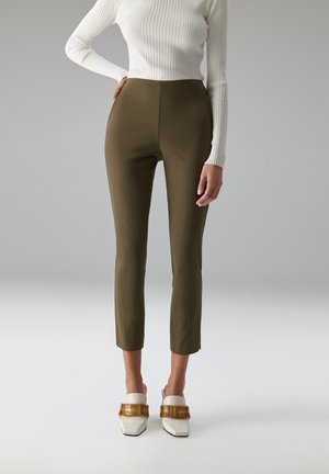 Frau trägt olivgrüne, eng anliegende, kurz geschnittene Hose, weißes, geripptes Langarmshirt und weiße Slipper mit goldener Kettendetail.