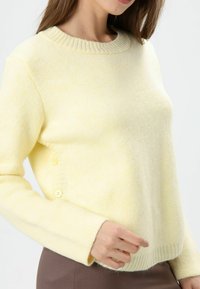 Gele gebreide trui met een geribbelde kraag, lange mouwen en knopen aan de zijkant. De stof heeft een zachte, gestructureerde uitstraling.