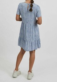 Kvinna som står med ryggen vänd framåt iförd en kortärmad blå mönstrad klänning och vita sneakers med rosa detaljer.