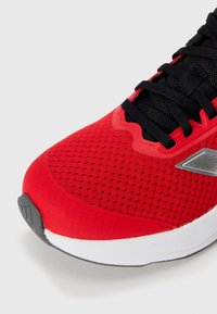Baskets de sport rouges avec un dessus en mesh, des lacets noirs et une semelle en caoutchouc grise. Les détails notables incluent un logo argenté et des panneaux texturés.