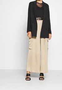 Svart skräddarsydd kavaj med dubbelknäppning, lager över en genomskinlig svart topp. Beiga vida cargobyxor med sidfickor och svarta klacksandaler.