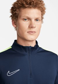 Giacca navy con zip e collo alto, con accenti verde lime sulle spalle e un logo Nike bianco sul petto. Texture liscia.