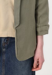 Grøn linnedjakke med opurgte ærmer, parret med en beige top og en mørkeblå denimnederdel med synlige sømme og lige snit.