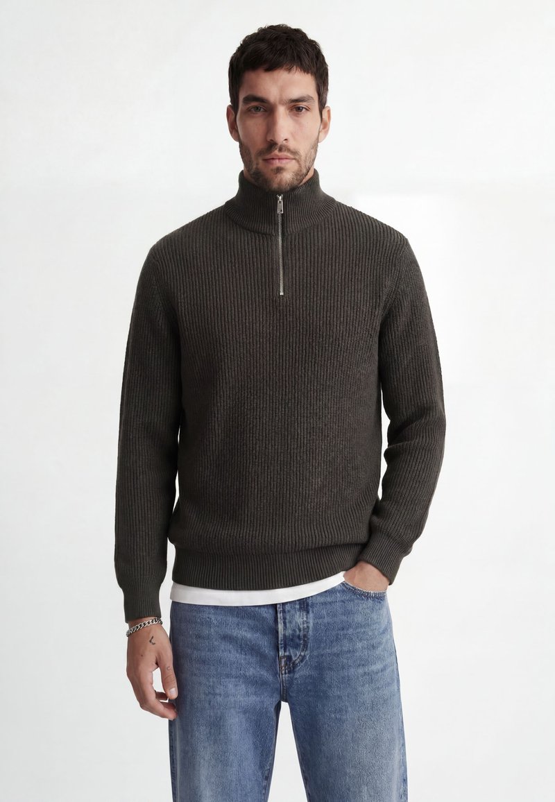 Homme portant un pull zippé côtelé vert foncé et un jean bleu, debout devant un fond clair uni.