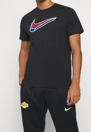 Hombre con camiseta negra de Nike con el logo Swoosh en patrón rojo y azul, y pantalones deportivos negros de Los Angeles Lakers con logotipos en la pierna.