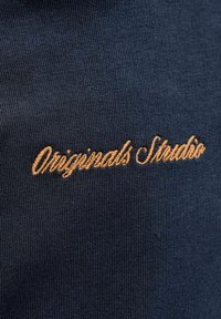 Tissu bleu marine à texture fine avec le texte brodé "Originals Studio" en fil d'or, positionné en diagonale.