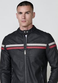 Uomo con capelli corti che indossa una giacca di pelle nera con strisce orizzontali bianche e rosse, in piedi contro uno sfondo chiaro e uniforme.