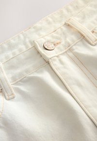 Gros plan d’un jean en denim couleur crème avec passants visibles, coutures et un bouton métallique étiqueté "Next Jeans Since 1982."