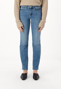 Narrow blue denim jeans dengan potongan mid-rise, dilengkapi dengan lima kantong, sedikit memudar, dan penutupan tombol. Dipakai dengan sweater rajut berwarna beige.