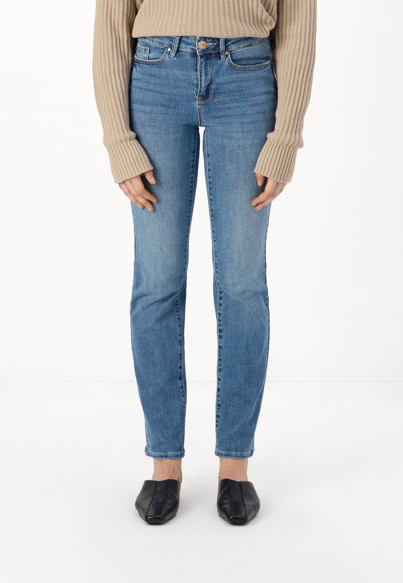 Narrow blue denim jeans dengan potongan mid-rise, dilengkapi dengan lima kantong, sedikit memudar, dan penutupan tombol. Dipakai dengan sweater rajut berwarna beige.