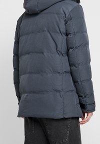 Chaqueta acolchada de color gris oscuro con capucha desmontable, diseño acolchado, puños elásticos y corte a media longitud. Textura suave y lisa.