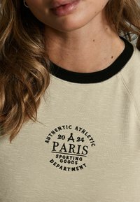 Heledbež sweatshirt musta ribiga kaelusega, millel on ümara kujuga graafikaprint, kus on tekst "PARIIS" ja "SPORDI KAUBAD". Pehme tekstuur.