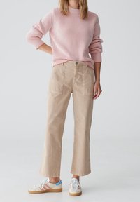 Pull en maille côtelée rose clair avec col montant, associé à un pantalon large en coton beige avec poches avant. Baskets blanches avec accents colorés.
