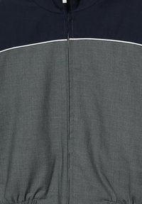 Veste avec une section supérieure bleu marine, une section inférieure grise, un détail de passepoil blanc au niveau de la poitrine, et une fermeture éclair centrale.