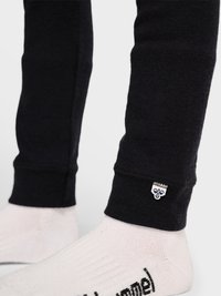 Underben iført sorte leggings med lille hvidt logo nær anklen, kombineret med hvide sokker med sort tekst på foden.