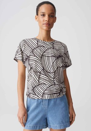 Donna che indossa una maglietta con motivo astratto bianco e nero e pantaloncini di jeans blu, in piedi davanti a uno sfondo chiaro uniforme.