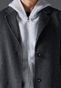 Grå uldfrakke med notch-lapel og to knapper, båret over en lysegrå zip-up hoodie. Teksturen i stoffet er synlig i begge beklædningsgenstande.