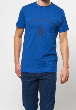 Homme portant un t-shirt bleu roi U.S. Polo Assn. avec un logo de joueur de polo et un jean bleu foncé, debout devant un fond uni.
