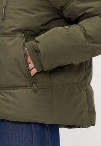 Hand deels in de ritssluiting van een olijfgroene gewatteerde jas met geribde manchetten over blauwe denim broek.