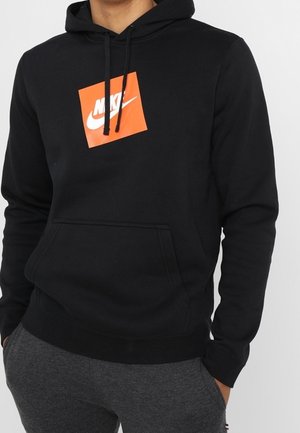 Osoba nosząca czarną bluzę z kapturem Nike z pomarańczowym kwadratowym logo i czarnymi troczkami, w zestawieniu z ciemnoszarymi spodniami, ręce w kieszeniach.