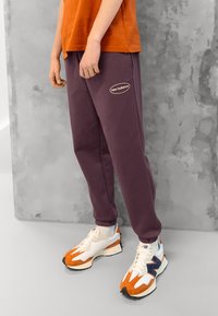 Fialové tepláky s elastickými manžetami a malým oválným logem "New Balance". Spárované s bílými a oranžovými teniskami s detailem z síťoviny a semiše.