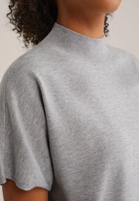 Světle šedý rolák vyrobený z měkké pleteniny, s těsným límcem a mírně oversized krátkými rukávy.
