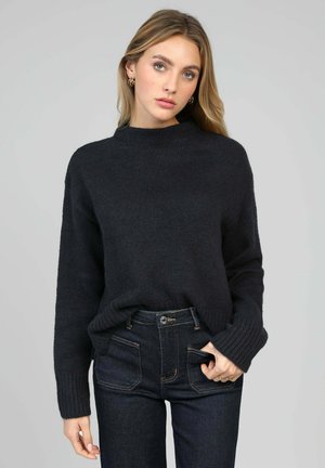 Femme aux longs cheveux blonds portant un pull en tricot noir et un jean foncé, debout devant un fond gris clair uni.