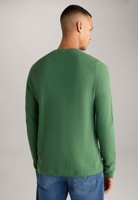 Groene lange mouwen shirt met een getextureerd gebreid patroon en een ronde hals, gecombineerd met blauwe jeans. Bevat een klein rood logo aan de zijkant.